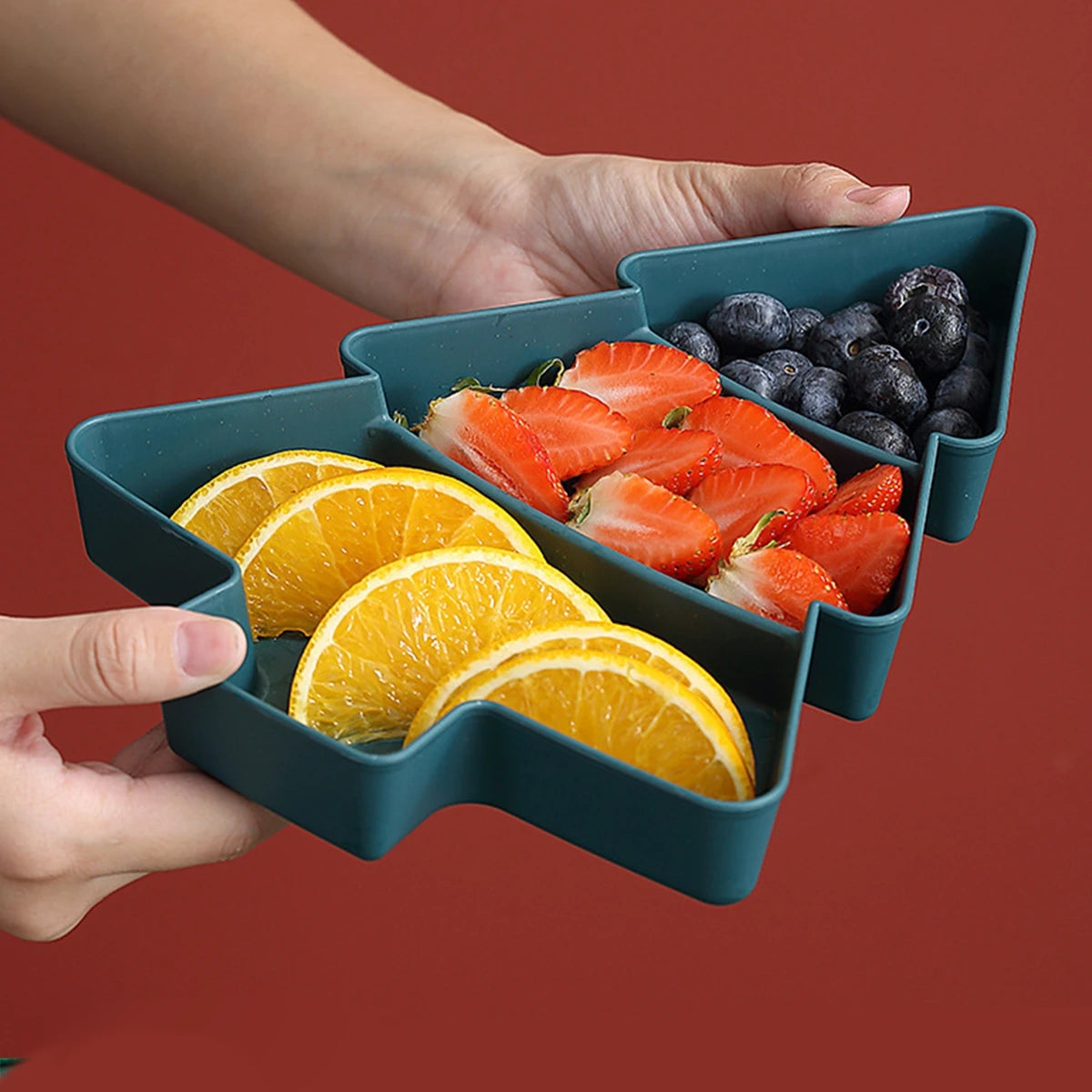 Hand holding a green compartmentalized container with sliced oranges and strawberries on a red background