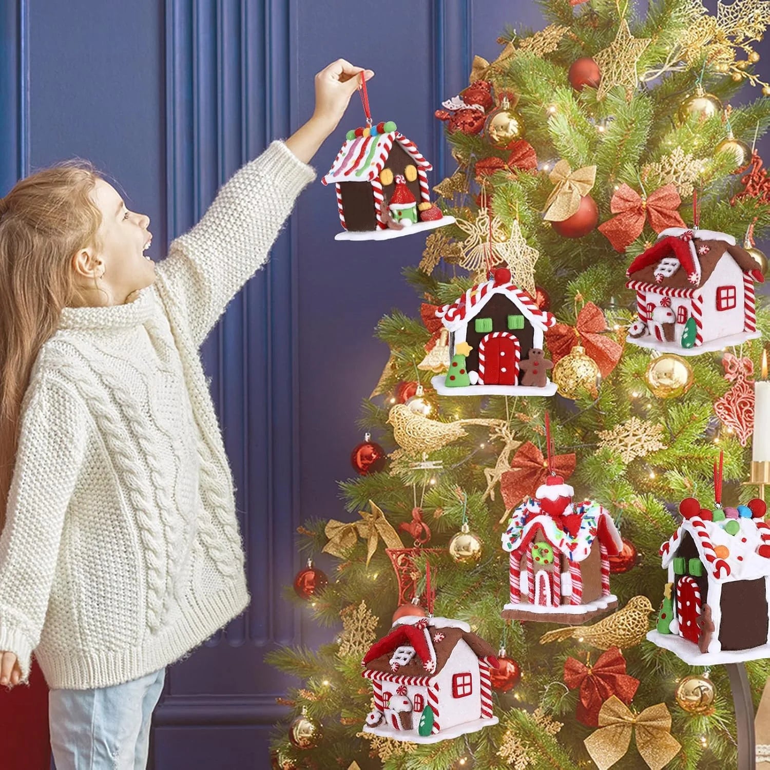 Child decorating a Christmas tree with gingerbread house ornaments against a blue wall.