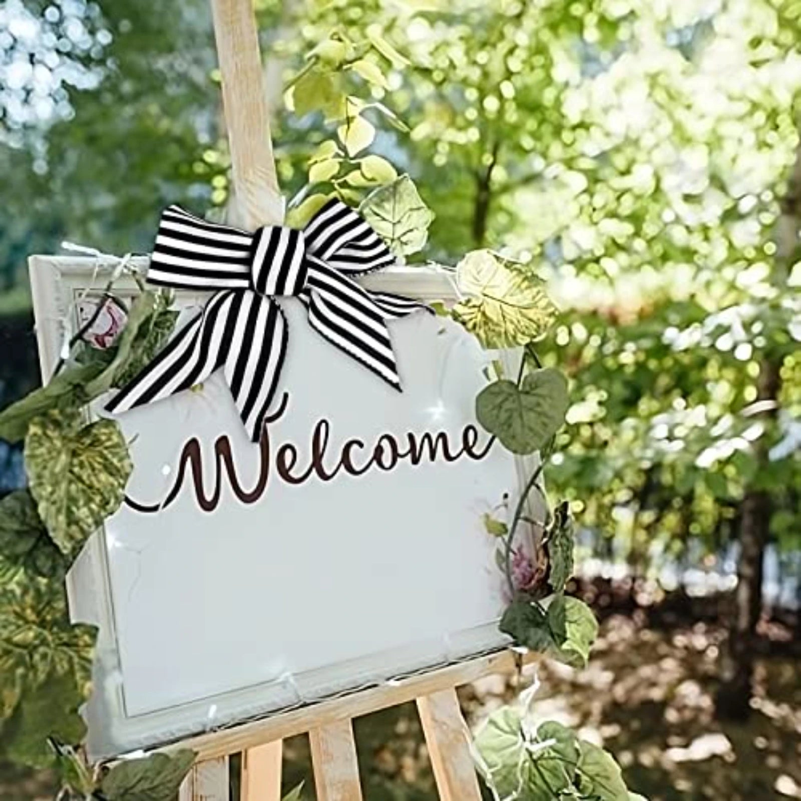 A rustic white wooden welcome sign on an easel, decorated with a large black and white striped bow, green leaves, and the word 'Welcome' in brown script, set against a blurred outdoor garden background.
