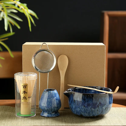 Tea-making set with blue ceramic pot, wooden spoon, metal strainer, and transparent container on a dark background.