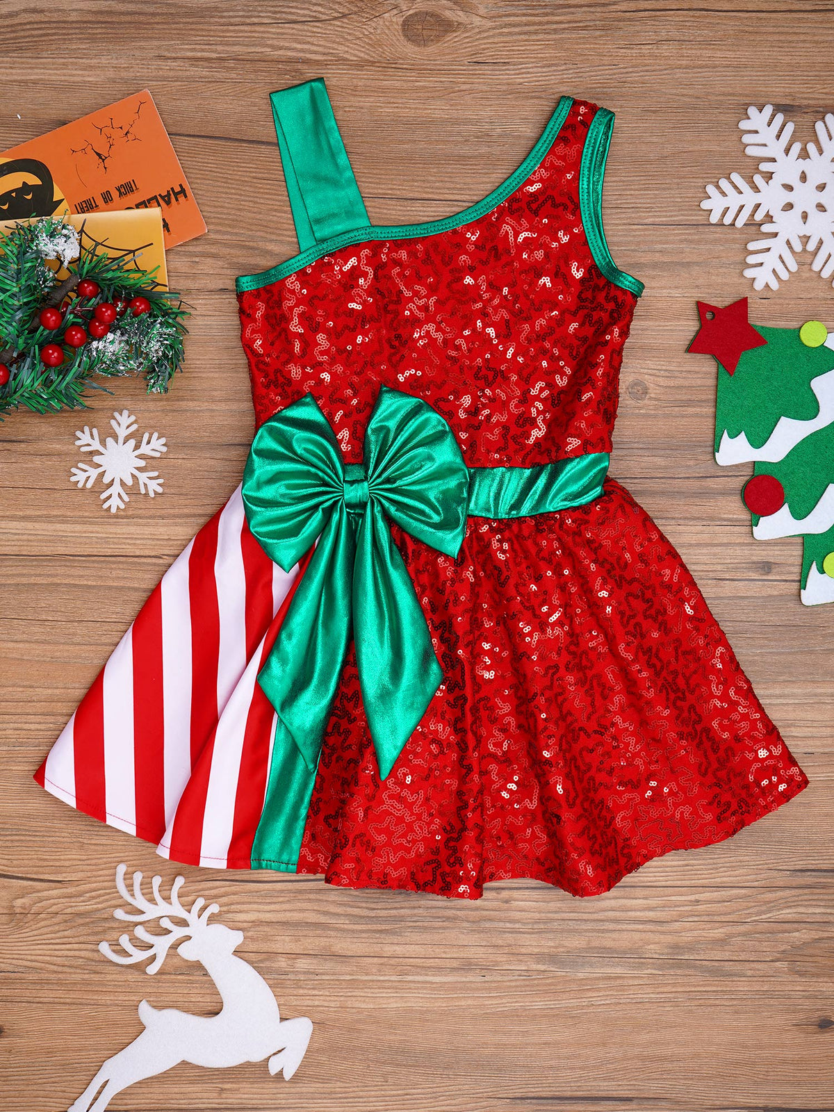 A festive Christmas-themed dress for girls, featuring a red sequined bodice with a large green satin bow at the waist, a green shoulder strap, and a red-and-white striped skirt panel. The dress is displayed in a flat-lay arrangement on a wooden background surrounded by holiday decorations.