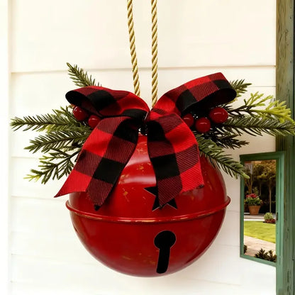 A red hanging metal bell decoration with a black keyhole cutout, adorned with a large red and black buffalo plaid bow, green pine branches, and red berries, hanging against a white exterior wall.