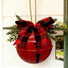 A red hanging metal bell decoration with a black keyhole cutout, adorned with a large red and black buffalo plaid bow, green pine branches, and red berries, hanging against a white exterior wall.