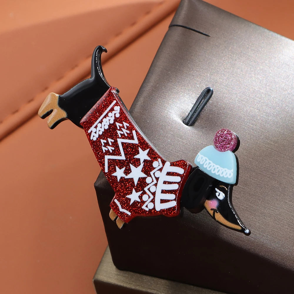 A dachshund-shaped enamel pin with a red glitter sweater featuring white star and zigzag patterns, wearing a light blue hat with a pink pom-pom, displayed on a dark metallic stand against a warm brown background.