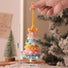 Handmade crocheted rainbow-colored tree ornament with measurements, held by a hand against a blurred indoor background.