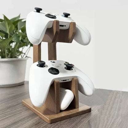 A wooden dual-controller charging stand holding two white wireless game controllers on a tabletop, with a blurred potted plant in the background.