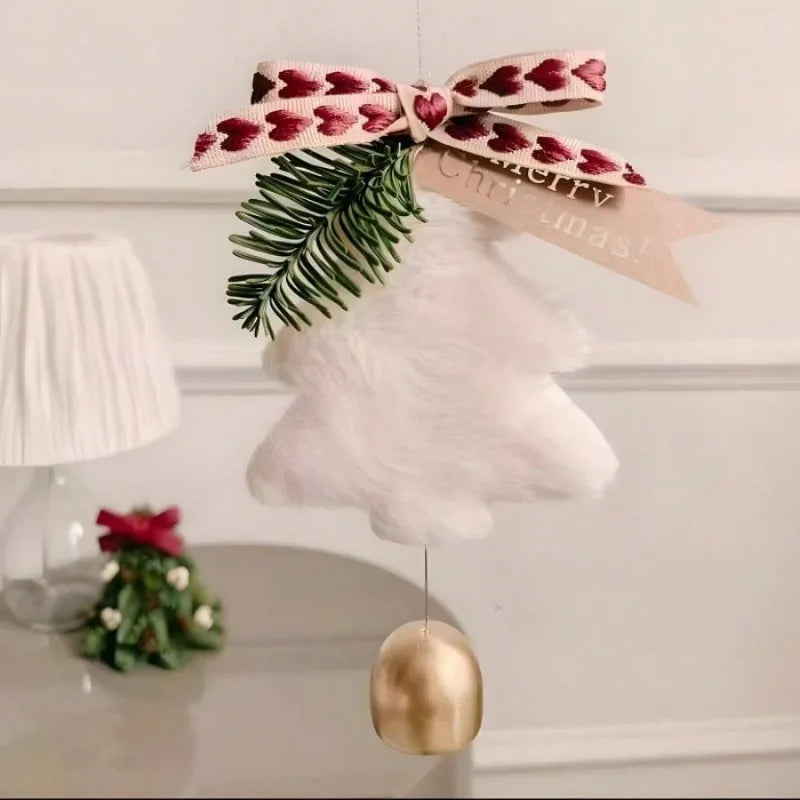 A fluffy white Christmas ornament shaped like a bell with a gold-colored hanging weight, decorated with a green pine branch and a pink ribbon with red hearts. A beige paper tag with 'Merry Christmas!' is attached.