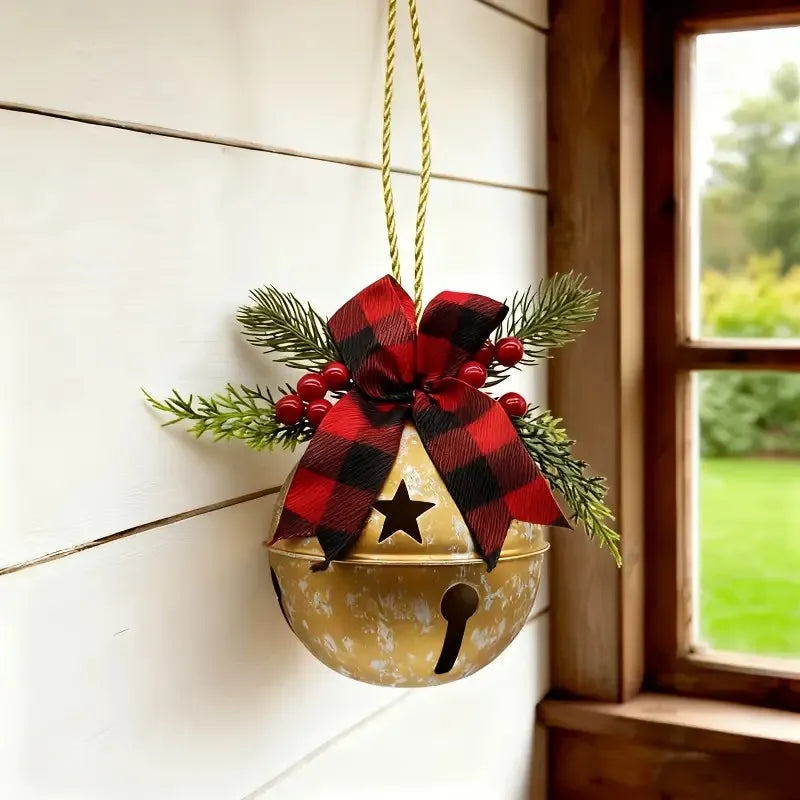 A rustic holiday hanging ornament featuring a small gold bell with a black bell hole, decorated with a red and black buffalo plaid ribbon bow, green pine branches, red berries, and a black star, hanging on a white wood paneled wall next to a window.