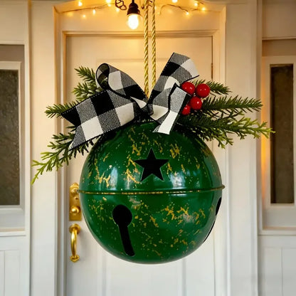 A green hanging Christmas bell decoration with gold distressed speckles, a black star cutout, a black handle, green pine branches, red berries, and a black-and-white buffalo plaid bow, hanging in front of a white door.