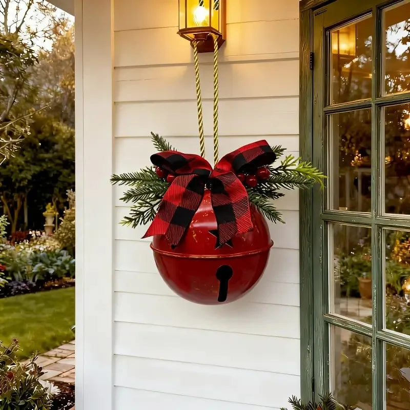 A red hanging decorative bell with a black bell-shaped detail, adorned with a red and black buffalo plaid bow and green pine branches, hanging on a white house exterior next to a green door.