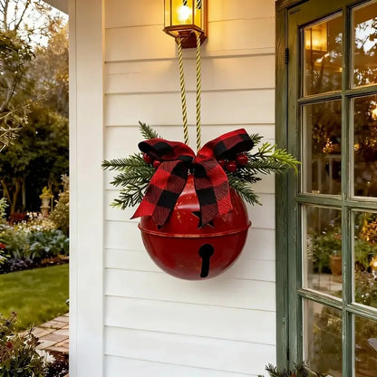 A red hanging decorative bell with a black bell-shaped detail, adorned with a red and black buffalo plaid bow and green pine branches, hanging on a white house exterior next to a green door.