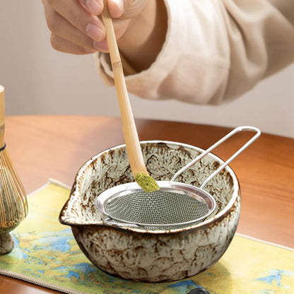 Person using a bamboo whisk to prepare tea in a ceramic bowl with a metal sieve.