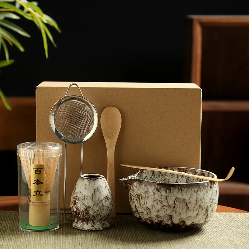 Tea-making set with a glass container, wooden spoon, and ceramic bowl on a green mat.