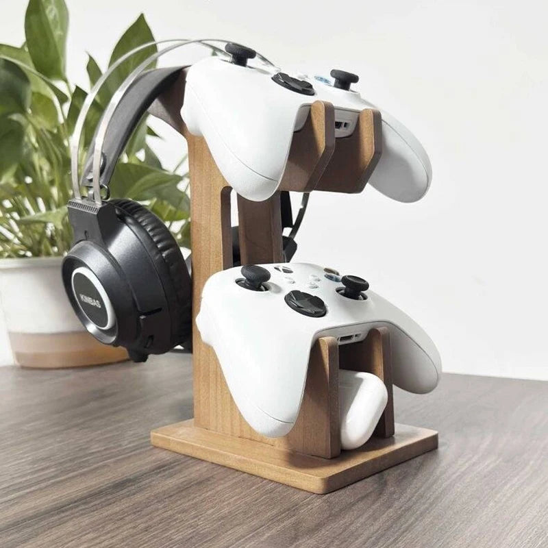 A wooden 3-in-1 game controller stand holding two white controllers and a pair of black over-ear headphones, placed on a wooden tabletop with a potted plant in the background.