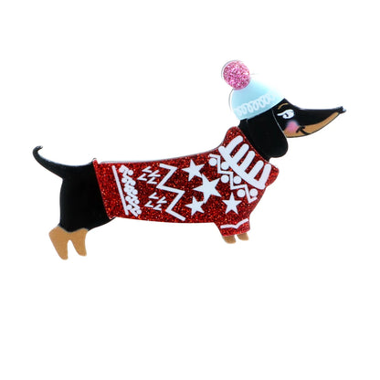 A dachshund-shaped enamel pin wearing a red glitter sweater with white snowflake patterns, a light blue beanie with a pink glitter pom-pom, and tan paws and snout, shown on a plain white background.