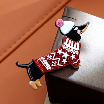 A dachshund-shaped enamel pin wearing a red glittery sweater with white geometric patterns, a white beanie with a pink pom-pom, and tan paws, resting on a dark brown box against a warm brown background.