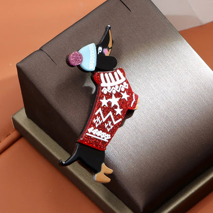 A dachshund-shaped holiday pin or brooch with a red glitter sweater, light blue hat, and pink pom-pom, displayed on a dark brown jewelry box against a warm tan background.