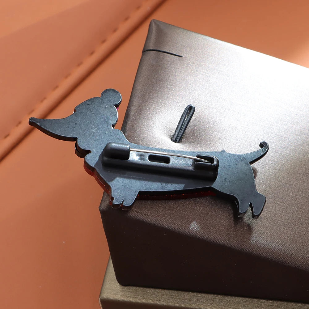 A black metal dachshund-shaped pin with a safety clasp, resting on a dark brown textured box against a warm brown background.