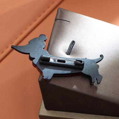 A black metal dachshund-shaped pin with a safety clasp, resting on a dark brown textured box against a warm brown background.